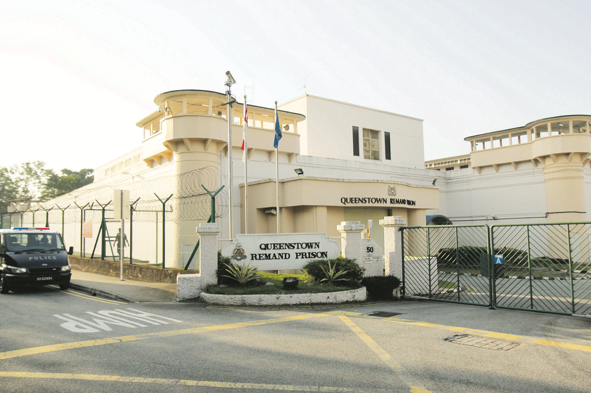 The image shows the entrance of Queenstown Remand Prison, with a police van parked outside and barbed wire fences visible.